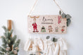 Load image into Gallery viewer, An der Wand hängendes nachhaltiges Adventskalenderschild mit 24 Adventsbeuteln. Motiv bunte weihnachtlich gekleidete Waldtiere. Dekomaterial Tannenzweige.