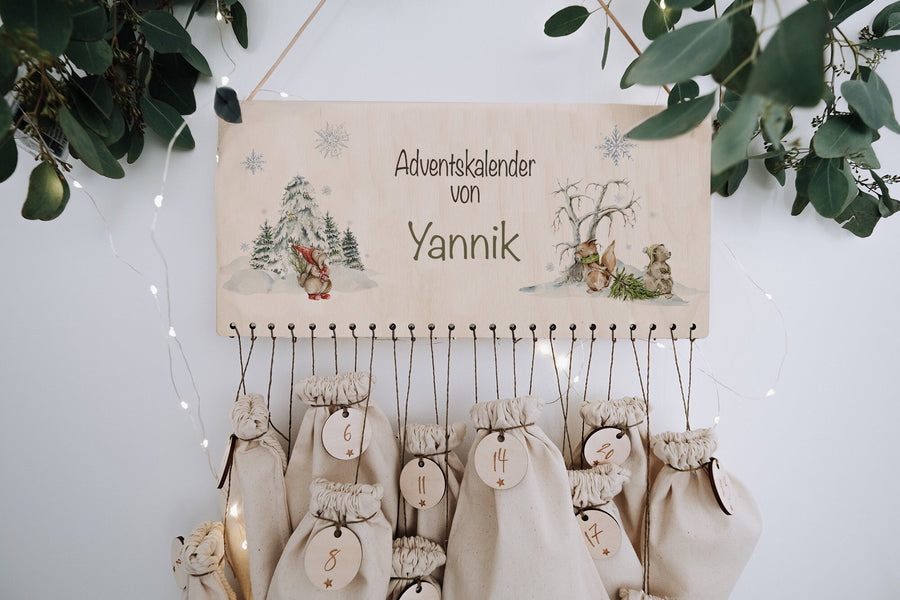 Adventskalender aus Holz, Schild bedruckt mit Winterwald, Eichhörnchen und Bär sowie Namen, darunter Stoffsäckchen