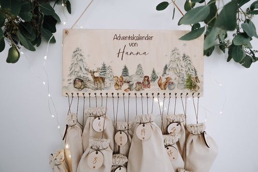 DIY Adventskalender als Schild, das an der wand hängt mit weihnachtlicher Winterlandschaft und Waldtieren. 24 mit Geschenken befüllte Jutebeutel hängen daran