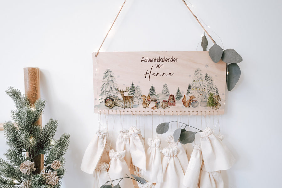 Adventskalender aus Holz mit Winterlandschaft und Waldtieren, mit Namen, darunter hängen helle Baumwollsäckchen